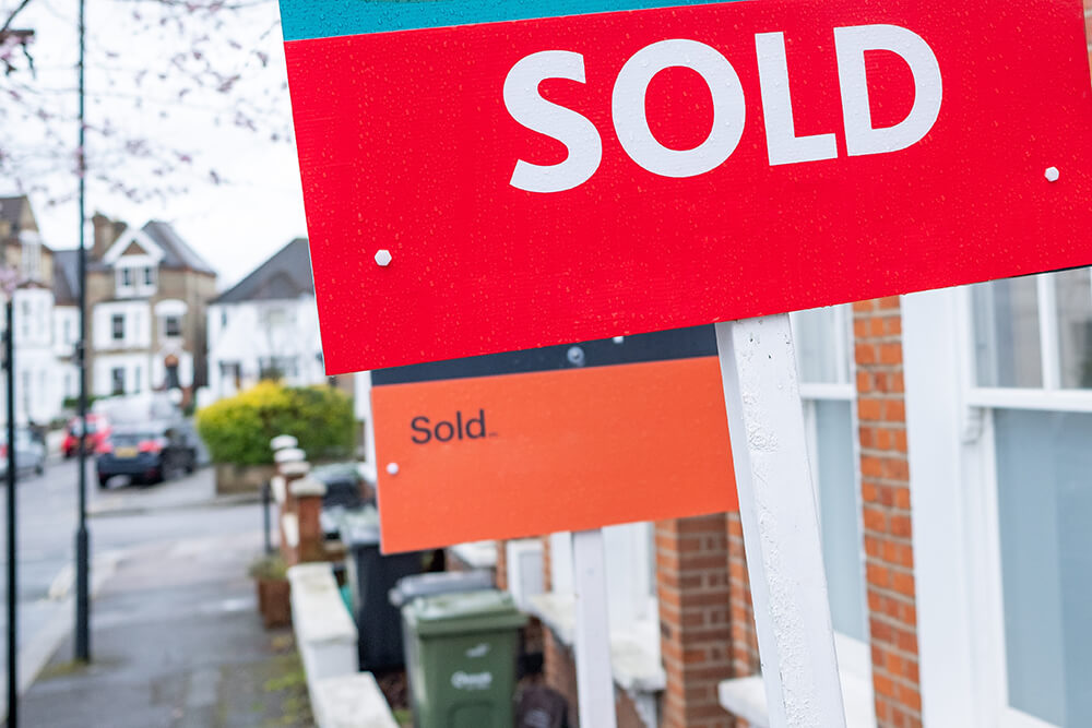 Prominent 'SOLD' estate agent sign in focus with a row of residential houses blurred in the background, symbolizing a swift and successful property sale.